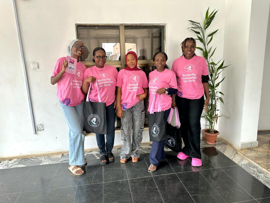 Five individuals wearing matching pink Butterfly Foundation for Girls t-shirts with white text stand indoors in front of a window and a potted plant. They are posing for a group photo on a tiled floor, with some holding black bags.