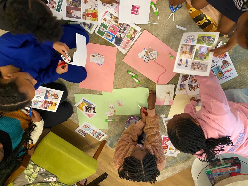 A group of children seated on the floor engaged in a collage-making activity. They are cutting out images from printed sheets and gluing them onto colored construction paper, surrounded by scissors, glue sticks, and craft materials.