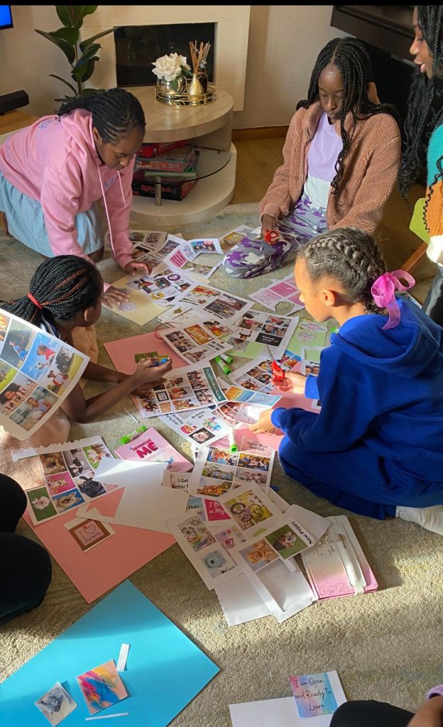 group of children sit on the floor in a cozy living room, engaged in an arts and crafts activity. They are surrounded by colorful papers, magazines, and cut-out images, working together on a collage or scrapbook project.