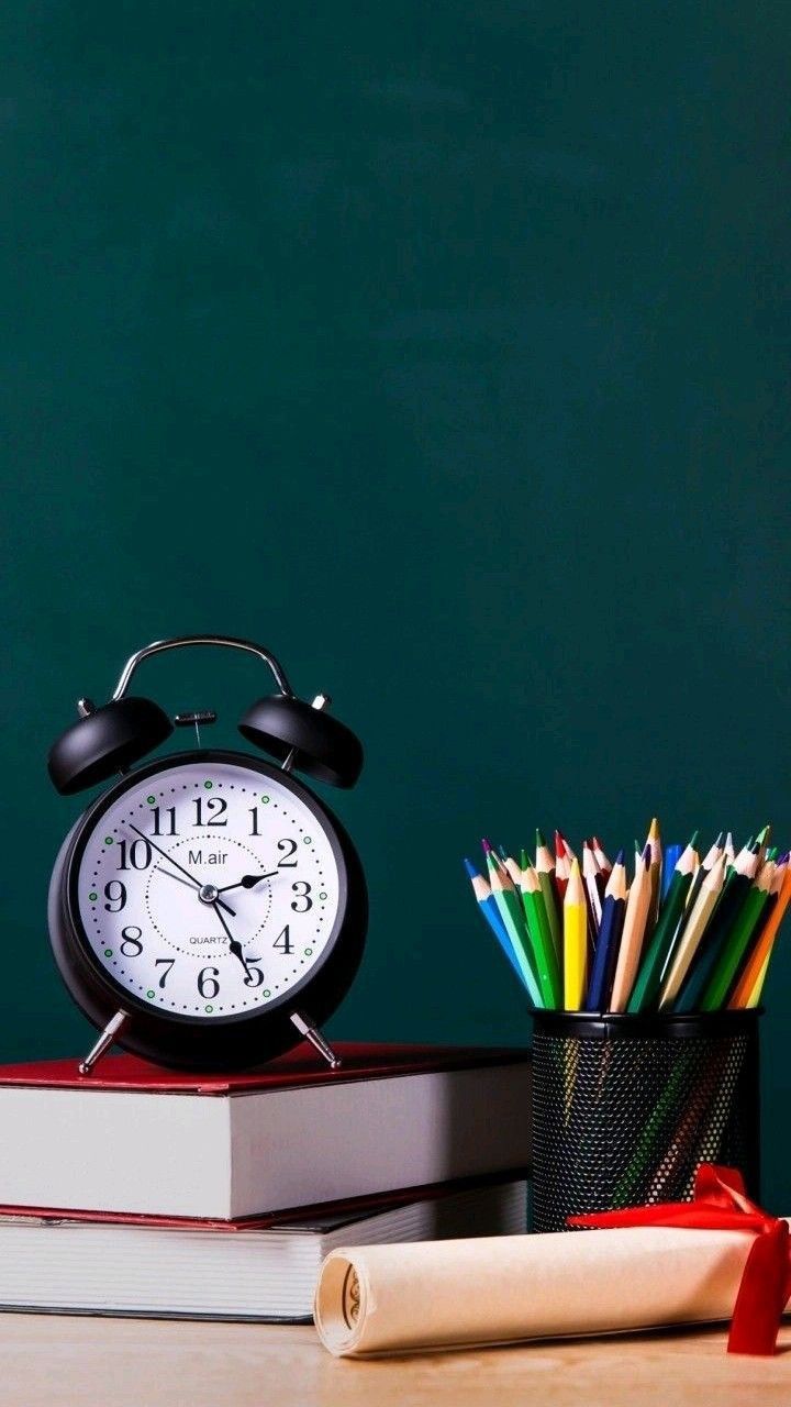 A black analog alarm clock sits atop two stacked red hardcover books next to a black mesh pencil holder filled with colorful pencils. A rolled-up parchment lies in front of the books, all set against a dark green chalkboard background, creating a cozy and academic atmosphere.