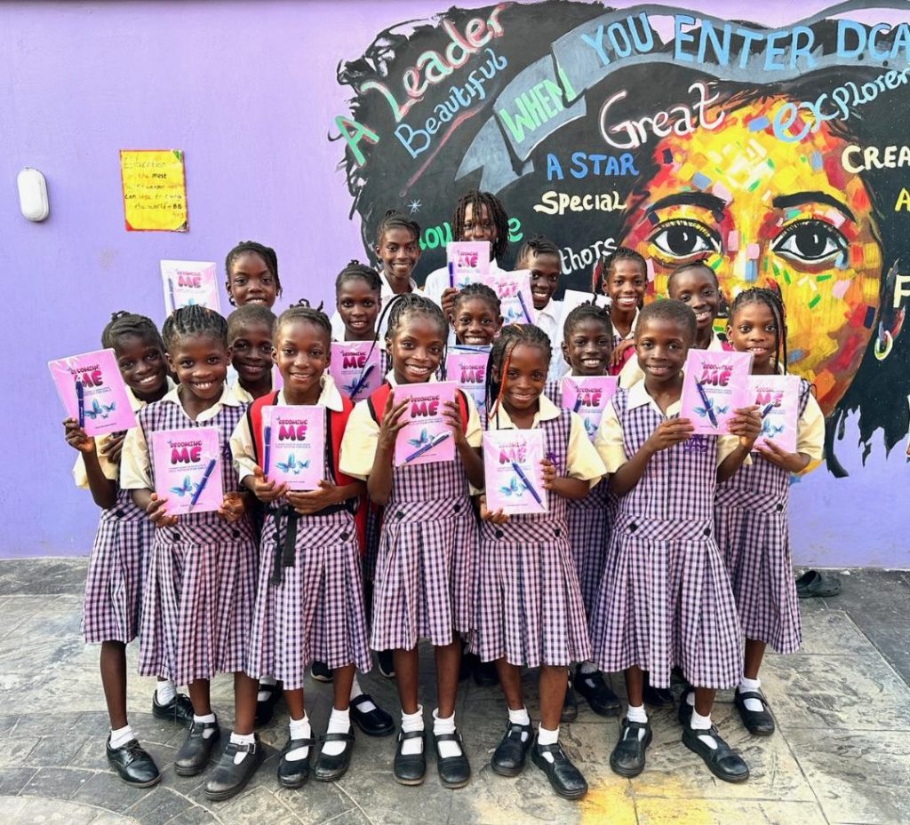 A group of schoolchildren in matching yellow shirts and purple plaid uniforms stand in front of a vibrant mural painted on a purple wall. The mural features a large, colorful face surrounded by uplifting words like 'Leader,' 'Creative,' 'Smart,' and 'Confident.' Each child holds a pink or purple folder, suggesting a school project or presentation. The scene conveys a joyful and empowering educational environment."