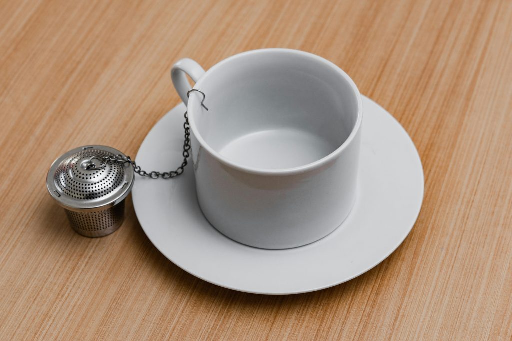 Close-up of a white ceramic cup on a saucer with a metallic tea infuser on wooden surface.