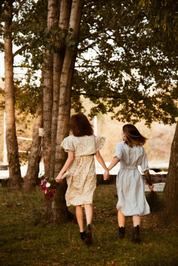 Two women in dresses hold hands as they walk through a sunlit forest path.