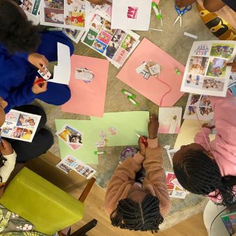 A group of children seated on the floor engaged in a collage-making activity. They are cutting out images from printed sheets and gluing them onto colored construction paper, surrounded by scissors, glue sticks, and craft materials.