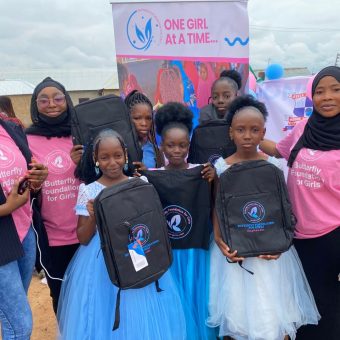 Group of young girls in blue and white dresses and women in pink shirts with black headscarves posing outdoors. Some hold black backpacks with logos. Behind them is a banner reading 'ONE GIRL AT A TIME...' with a stylized girl and wings. The scene suggests a community event focused on empowering girls."