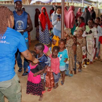 Children and adults stand in a line inside a building, receiving aid or services from individuals in blue shirts. One shirt displays the text 'UNHCR' and 'Welcome...', indicating involvement from the United Nations High Commissioner for Refugees. The setting resembles a humanitarian aid or registration event.