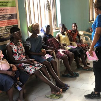 Seven people sit on plastic chairs in a waiting area while one person stands holding a pink booklet. The room has tiled flooring, a window with blinds, and a health-related poster on the wall.