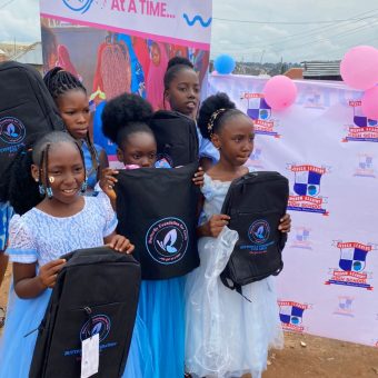 Five children dressed in formal or semi-formal clothing stand in front of a decorated backdrop holding black backpacks with a stylized bird logo. The backdrop features promotional material with anatomical heart images and is surrounded by pink and blue balloons.