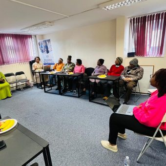 Nine people sit in a row along a wall in a classroom or meeting room, with one person in a bright pink top seated separately. The room has white walls, blue carpet, and purple blinds.