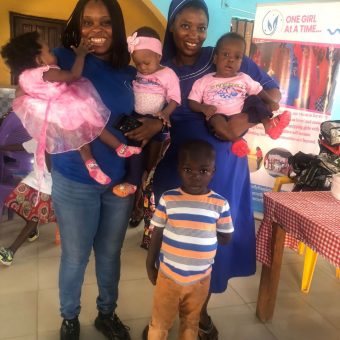 Girls in blue dresses and women in pink shirts pose with backpacks near a banner reading 'ONE GIRL AT A TIME...'.