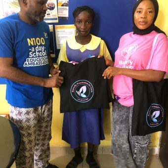 Three individuals standing indoors. The person on the left wears a blue shirt with yellow text reading 'N100 a day School Support Abroad.' The middle person, a young girl in a yellow and blue school uniform, and the person on the right, in a pink shirt and patterned pants, both hold black tote bags with the butterfly foundation for girls logo.